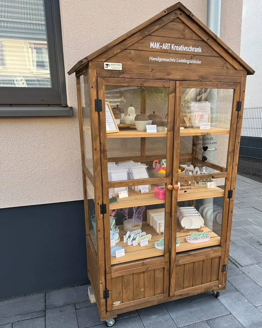 Der MAK-ART Kreativschrank: ein Holzschrank mit Giebeldach und Glastüren an einer Hauswand, in den Regalen handgemachte Deko und Karten