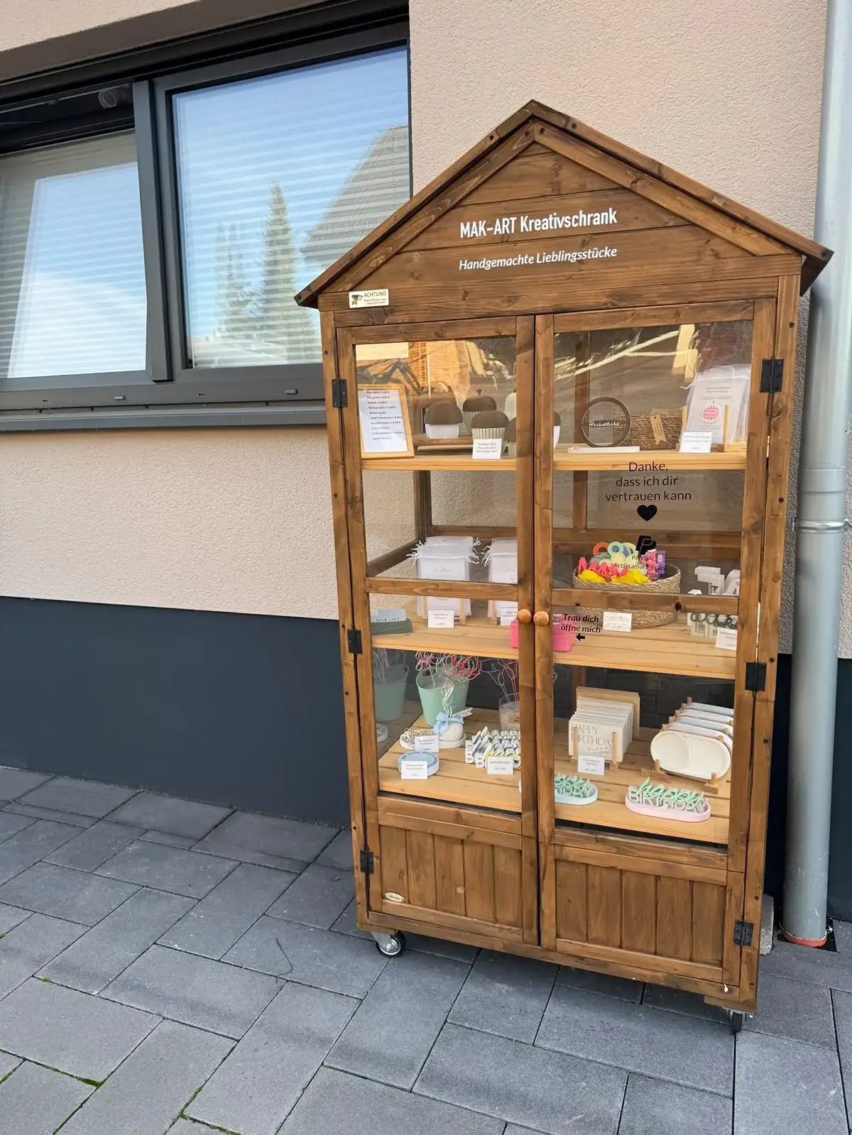 Der MAK-ART Kreativschrank am Talweg 54: ein Holzschrank mit Giebeldach und Glastüren vor einer Hauswand, darin handgemachte Deko, Karten und Schriftzüge